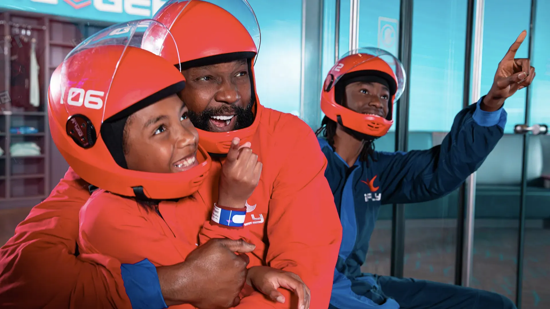 Family in red and blue iFLY flight suits smiling and pointing toward the indoor skydiving wind tunnel.